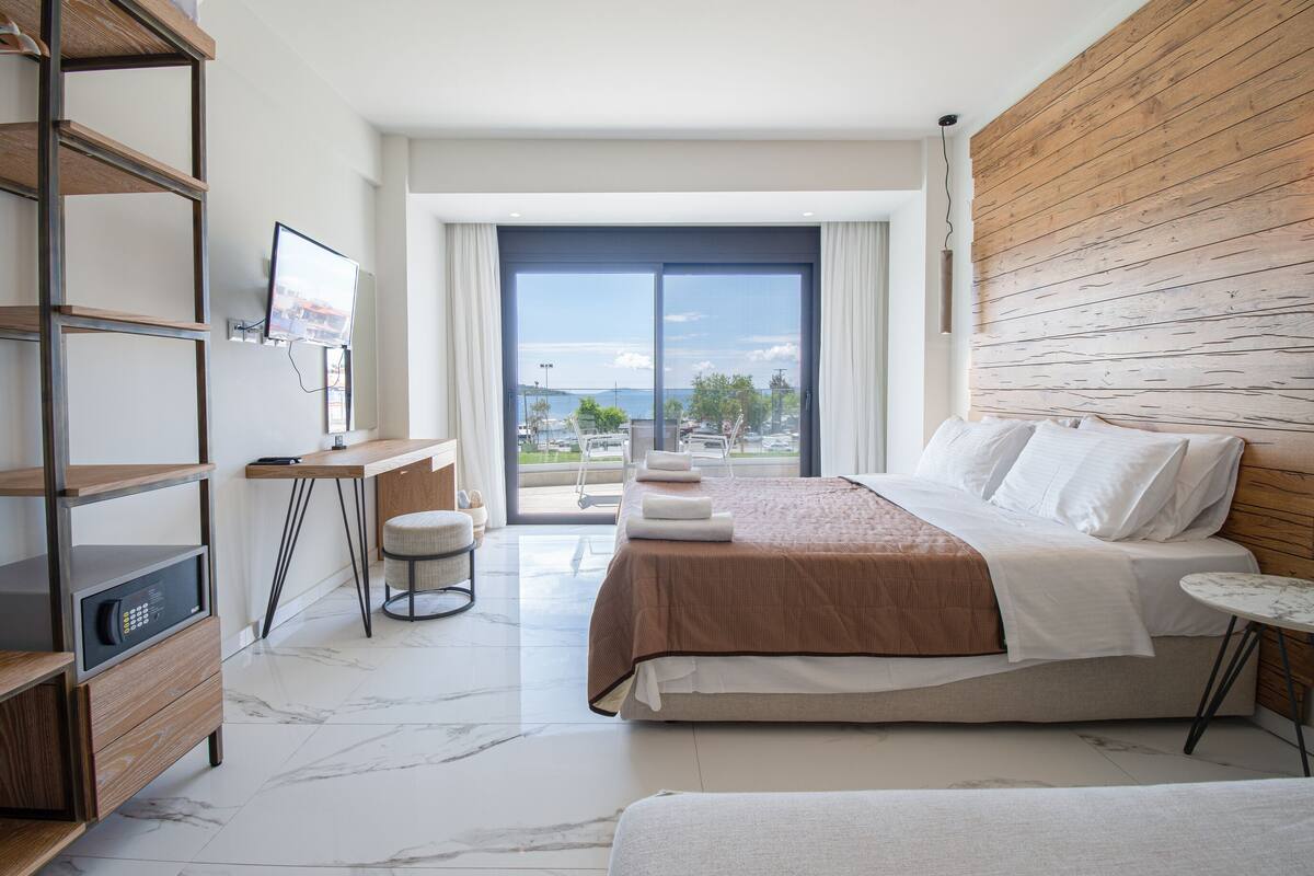A modern hotel room with a large bed, a desk with a TV, and a view of the sea.