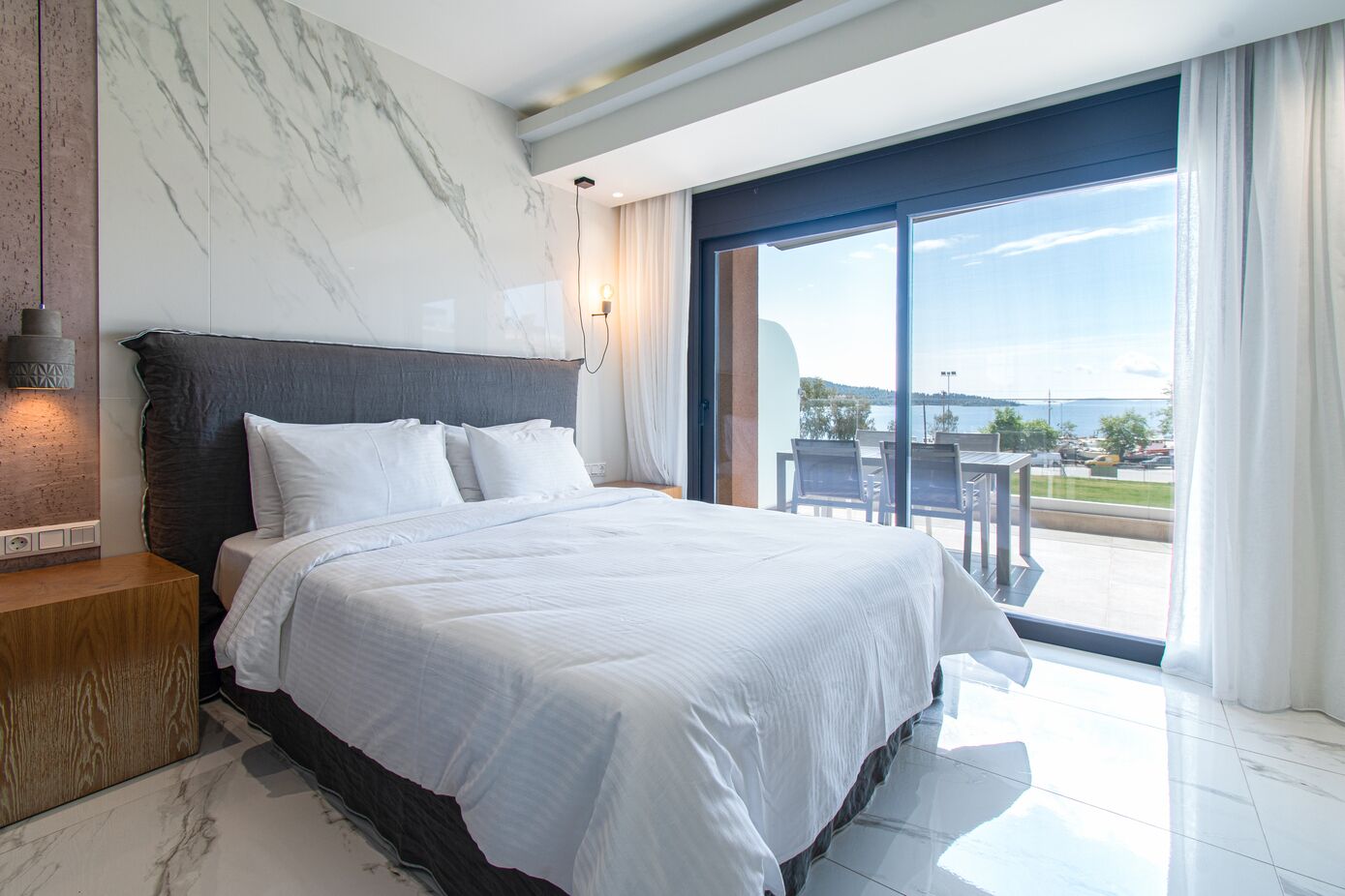 Une chambre à coucher moderne avec un grand lit, des murs en marbre et un balcon offrant une vue sur l’eau.