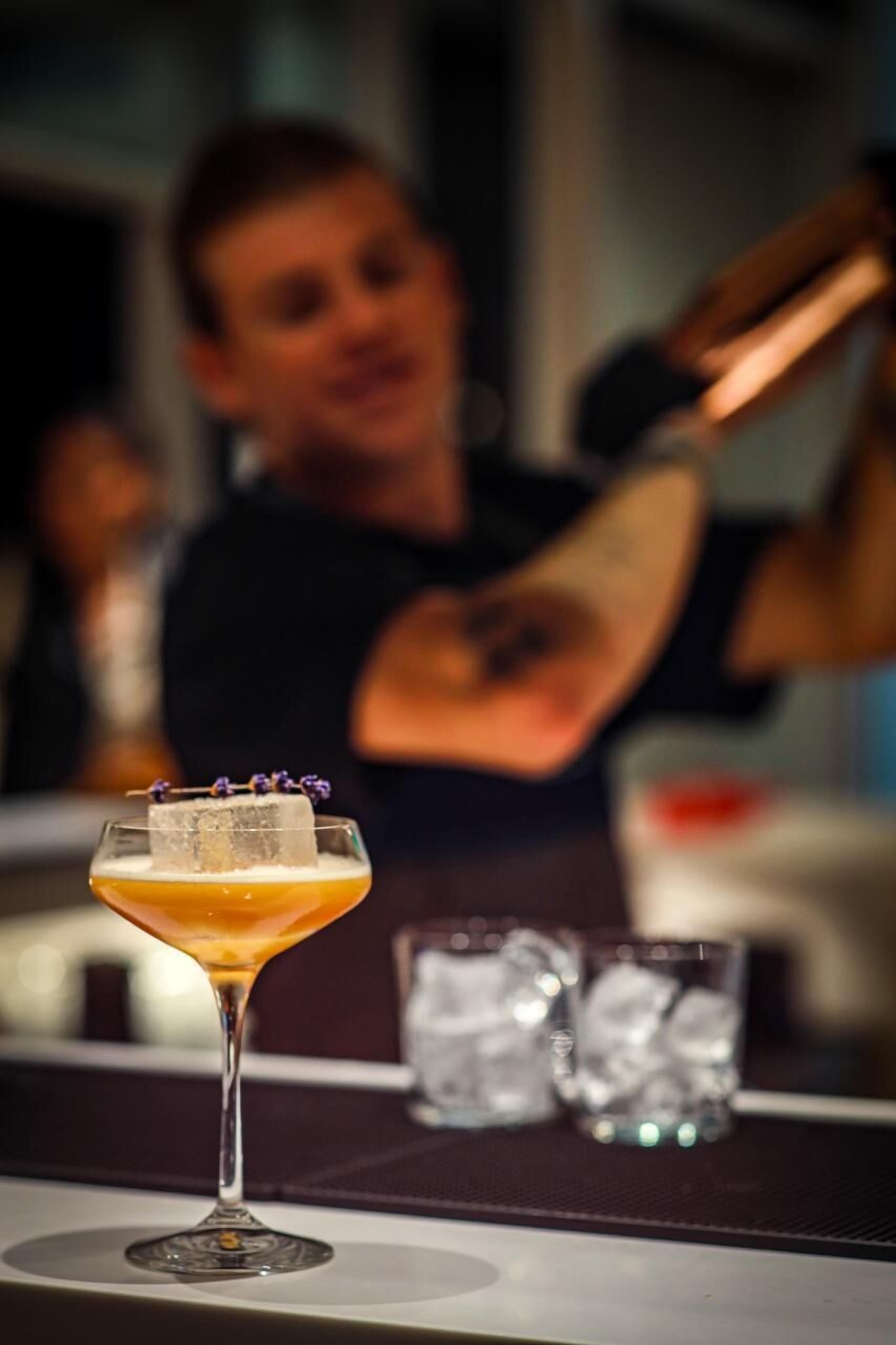 Un cocktail servi dans un verre à martini, garni d’une tranche d’orange et d’une fleur, avec des glaçons dans le verre et un arrière-plan flou représentant une personne.