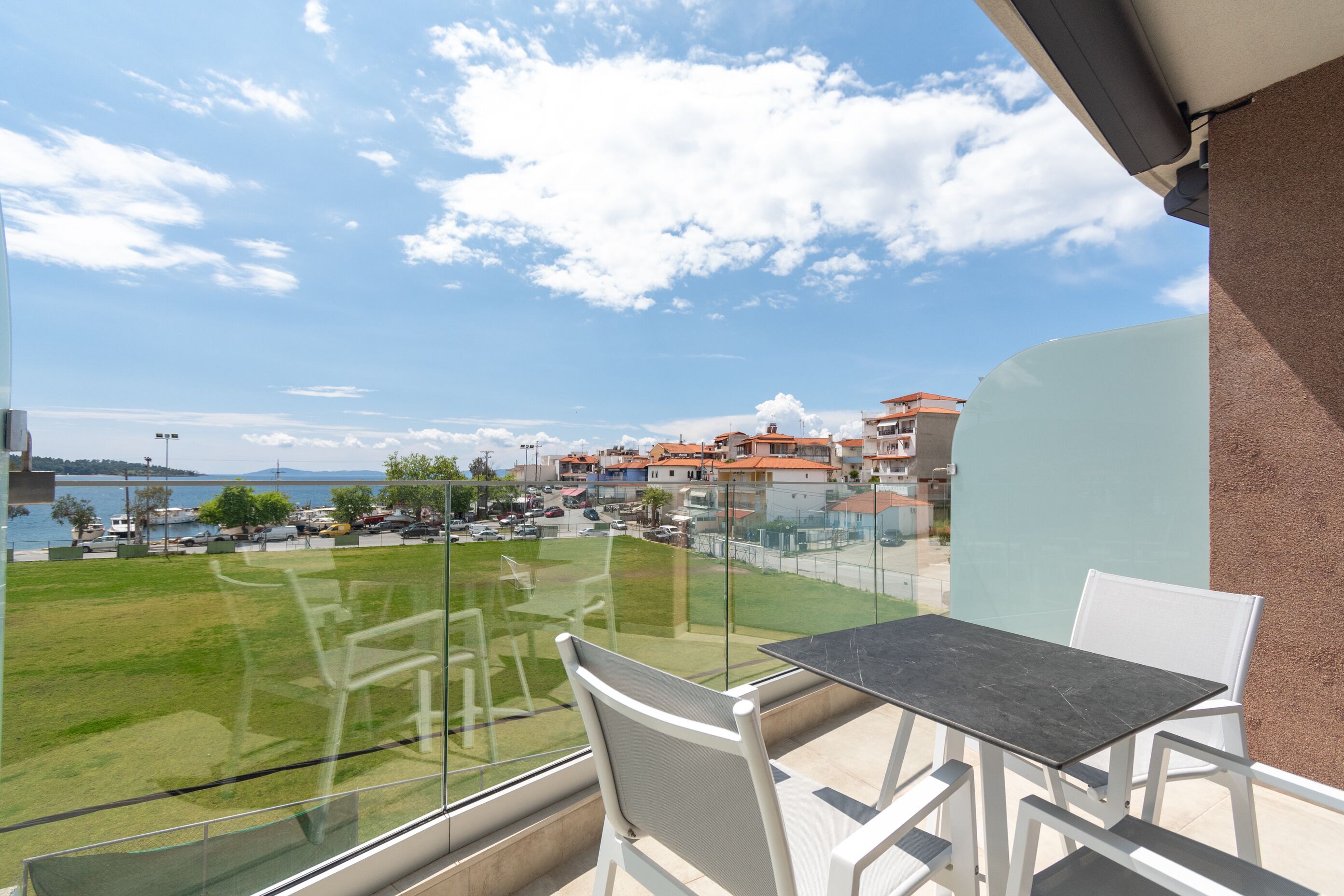 A balcony with a glass railing overlooking a grassy area and a body of water. There is a table and two chairs.