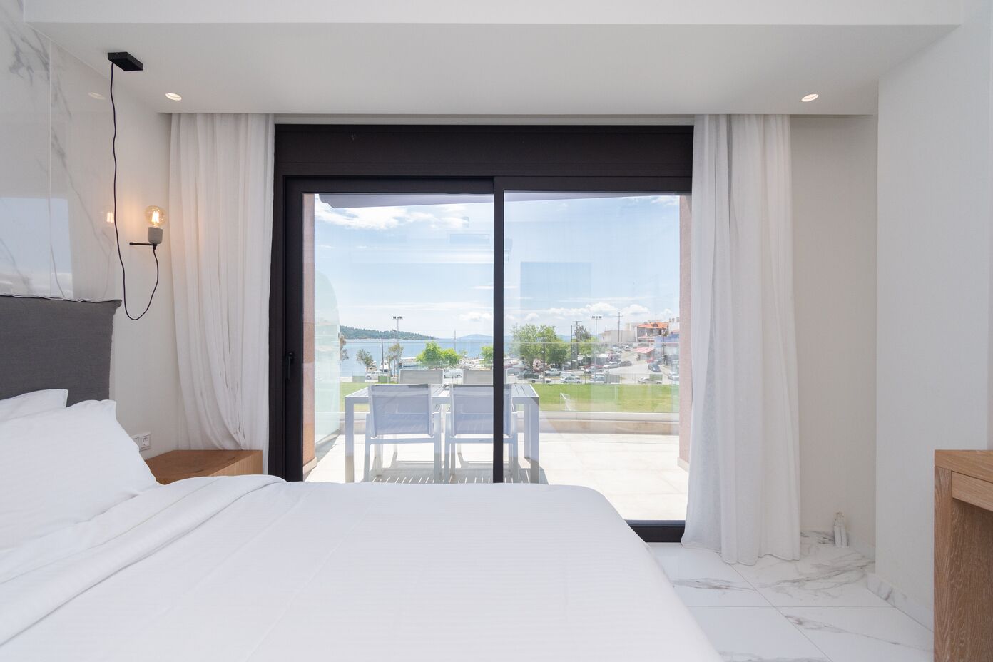 Habitación de hotel moderna con una cama grande, un balcón con mesa y sillas, y vistas a la ciudad y al agua.