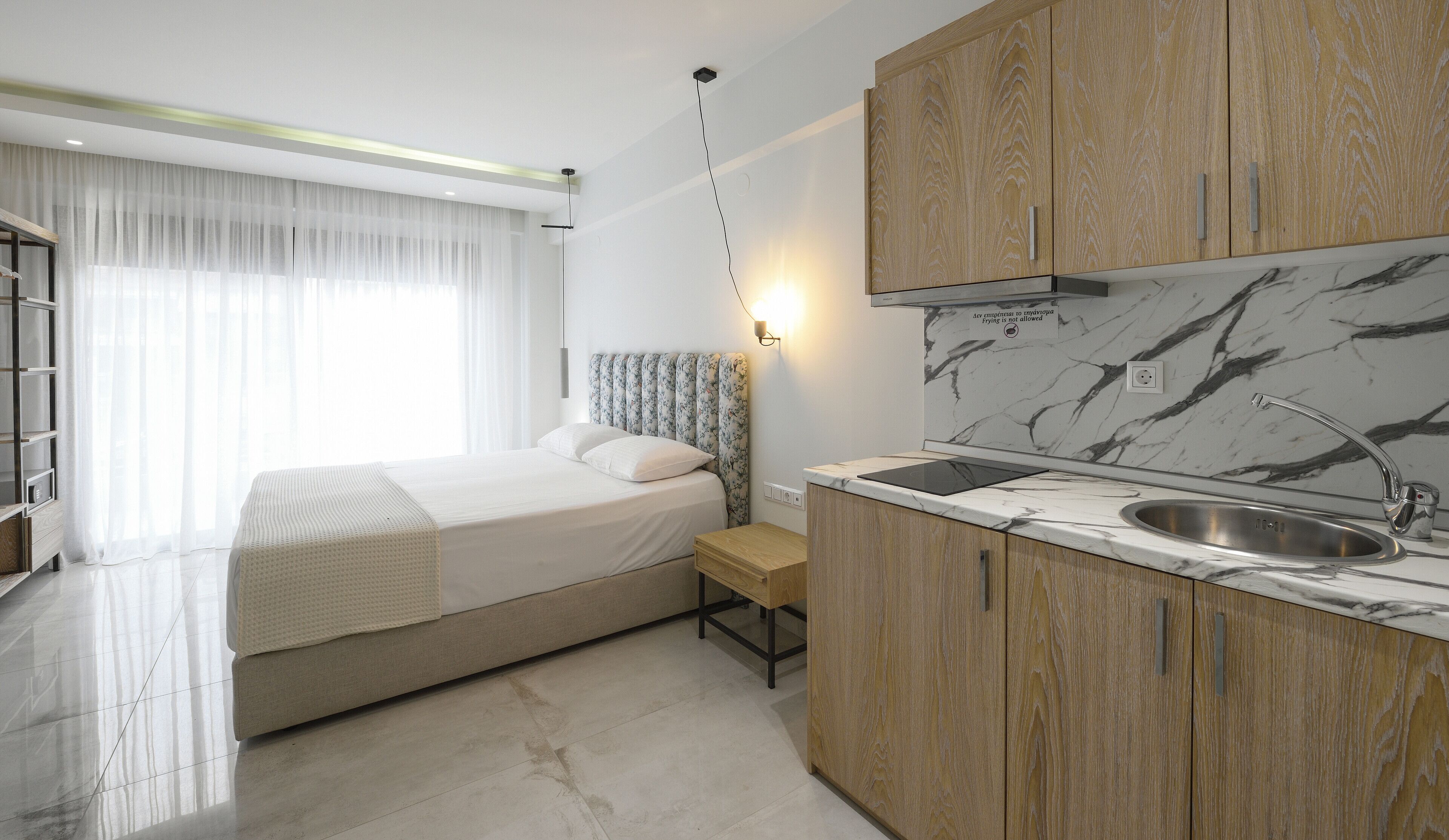 A modern hotel room with a bed, a small bedside table, a built-in bench, a kitchen area with a sink and countertop, and a large window with sheer curtains.