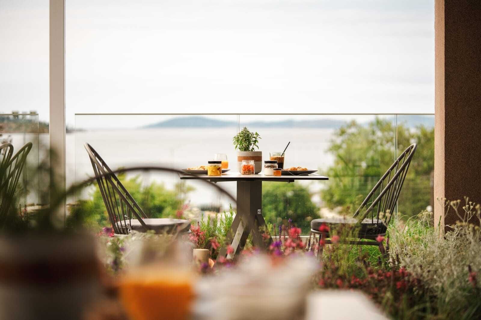 Une table à manger, dressée avec des plats et des boissons, entourée de chaises, avec une vue sur la mer et les montagnes en arrière-plan.