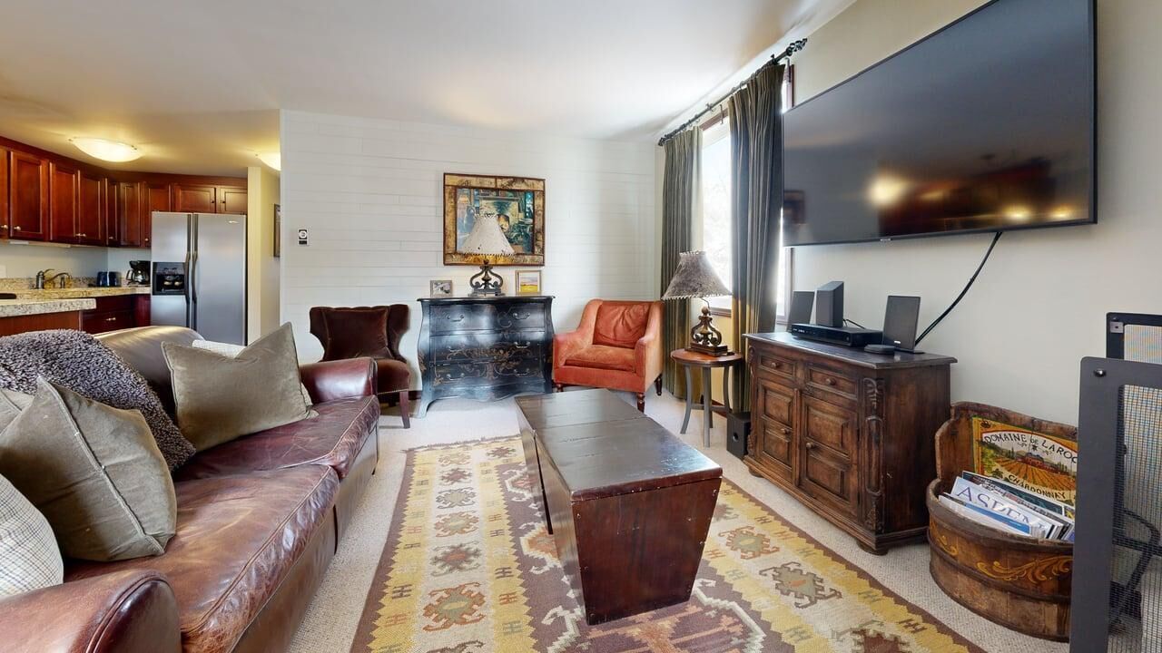 A living room with a sofa, coffee table, and a television. There is a kitchen area with cabinets and a refrigerator.