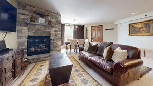 A living room with a stone fireplace, leather sofa, and a dining area with a chandelier. - Terracehouse 93 - Steps to Slopes | Inviting Mountain Retreat, Pool & Hot Tub (Snowmass Village)