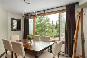 A dining room with a wooden table, six chairs, a chandelier, a mirror, and a view of trees through large windows. - Terracehouse 93 - Steps to Slopes | Inviting Mountain Retreat, Pool & Hot Tub (Snowmass Village)