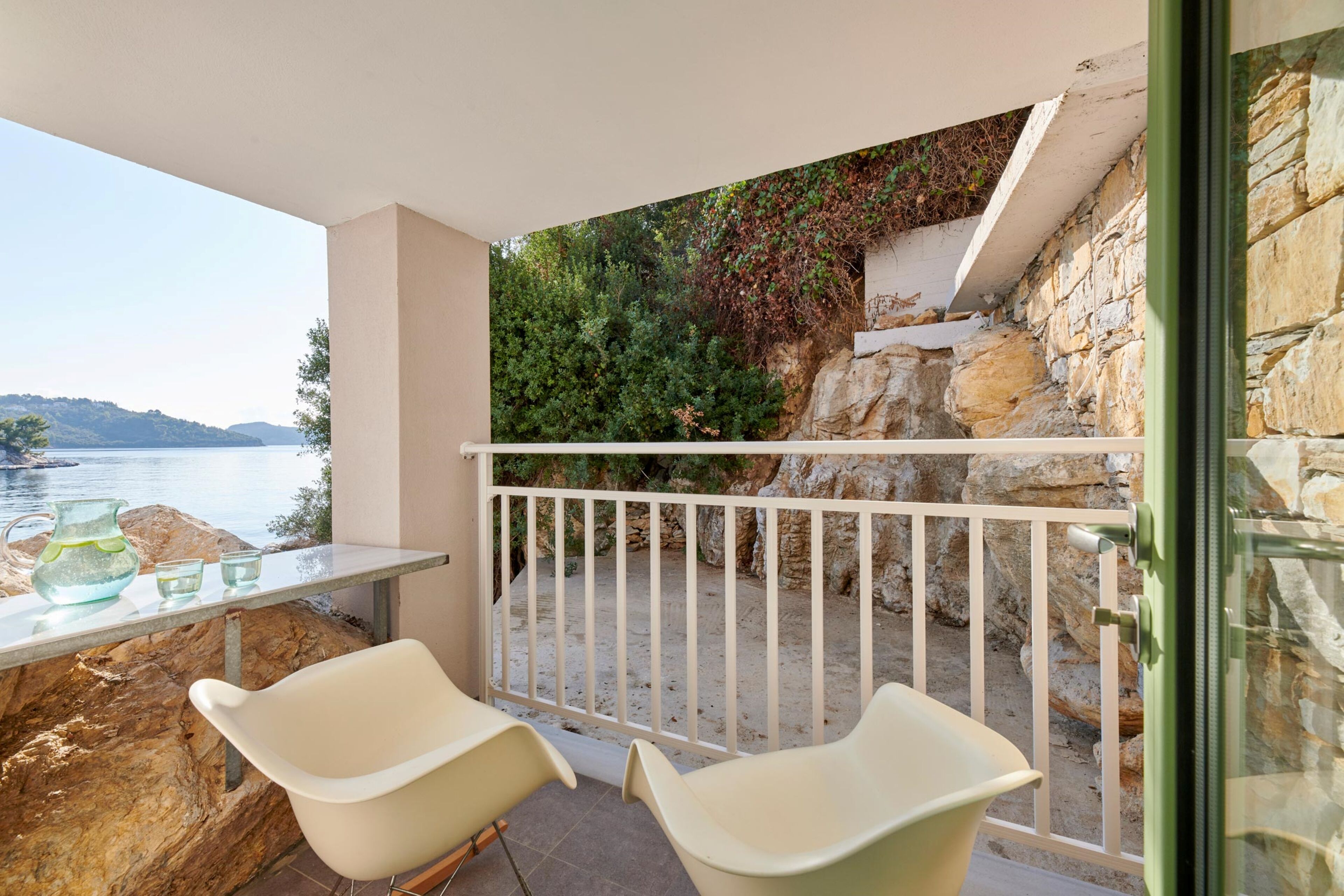 Un balcon avec vue sur la mer, deux chaises blanches et une table avec des verres et une carafe.
