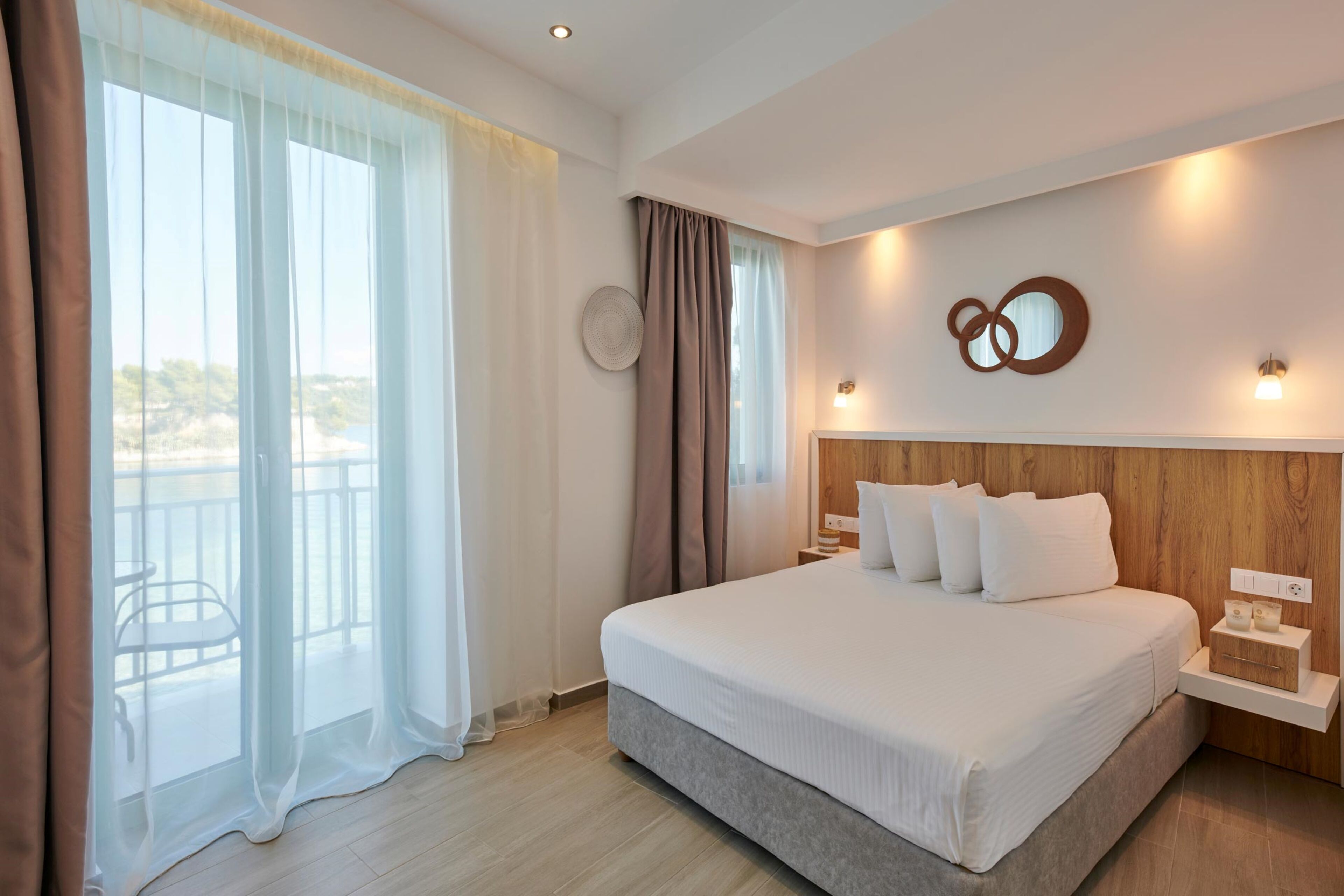 Une chambre d’hôtel moderne avec un grand lit, une tête de lit en bois, une table de chevet avec une lampe et une vue sur l’extérieur.