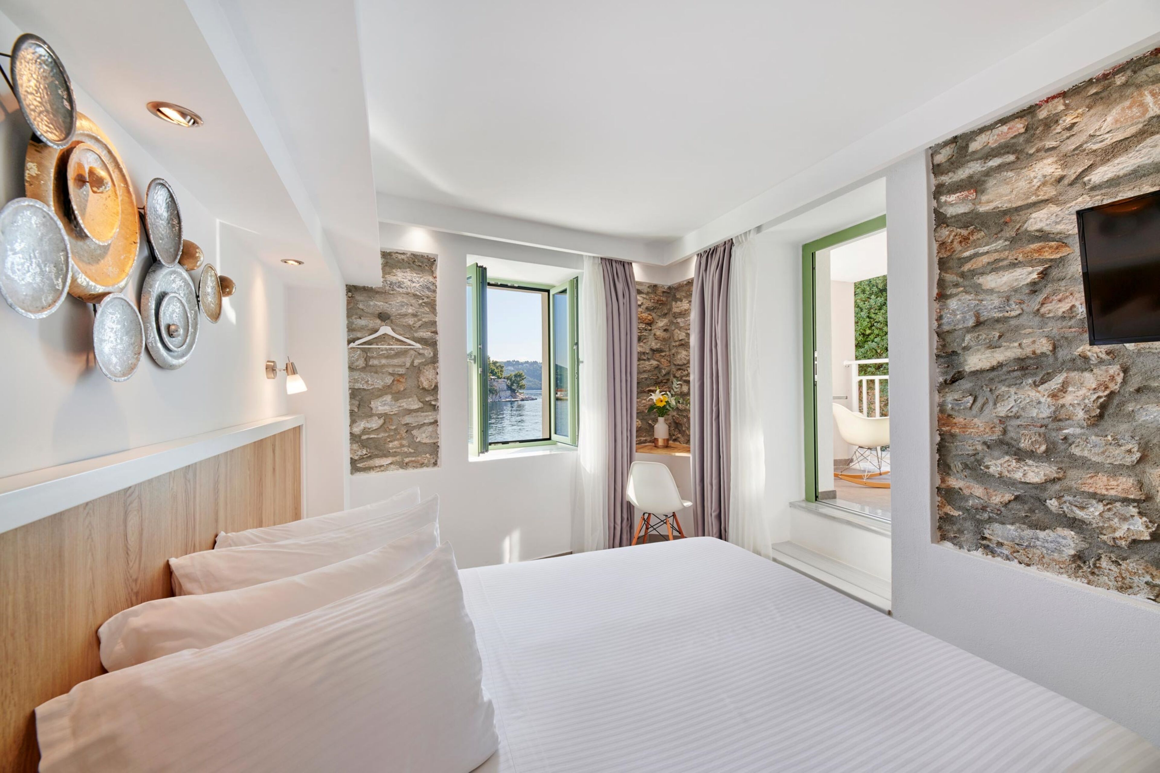 A modern hotel room with a large bed, a stone wall, a view of the outdoors, and a decorative metal wall art piece.