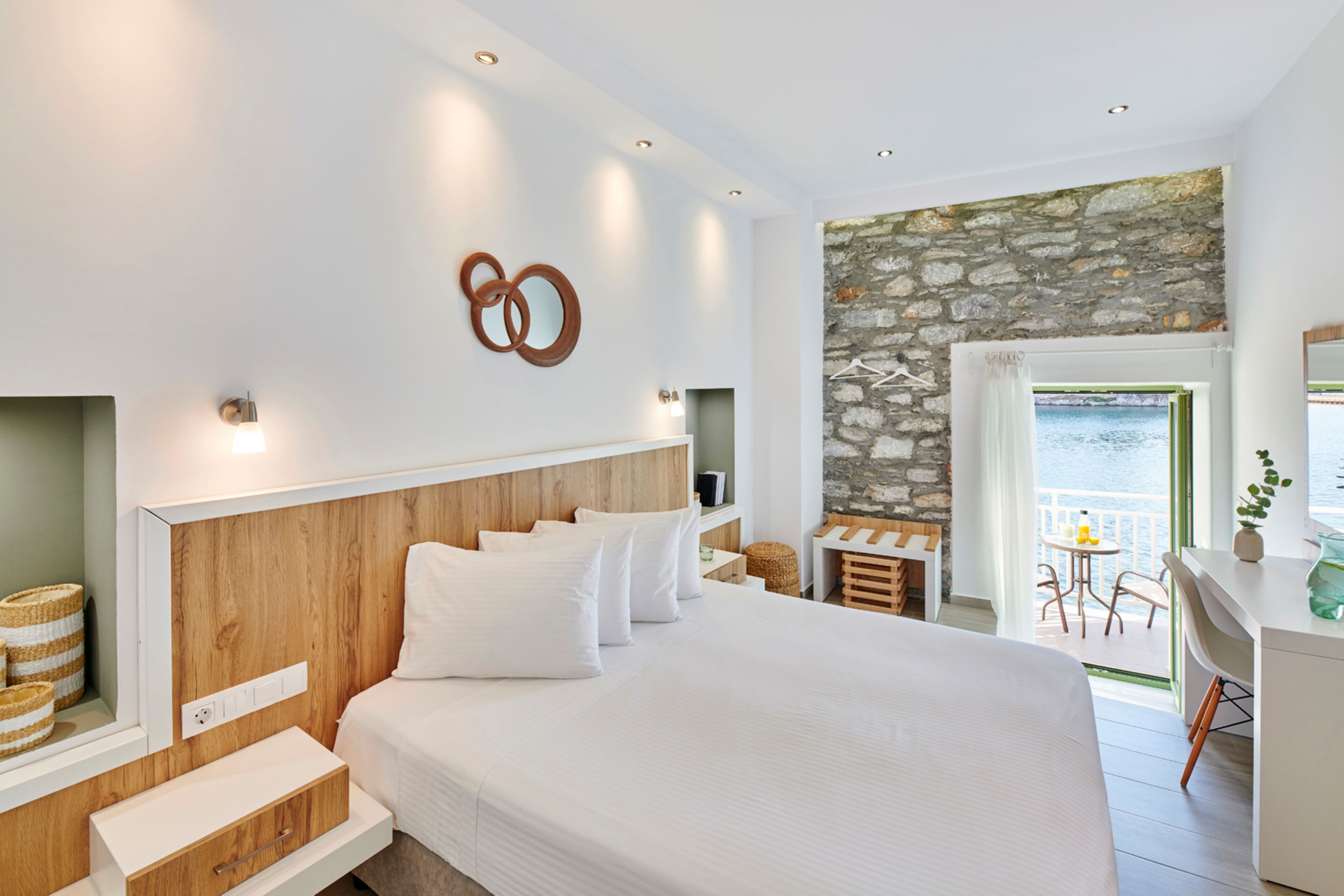 Un dormitorio moderno con una cama grande, cabecero de madera, ropa de cama blanca, pared de acento de piedra, un balcón con vistas y un escritorio con una planta.