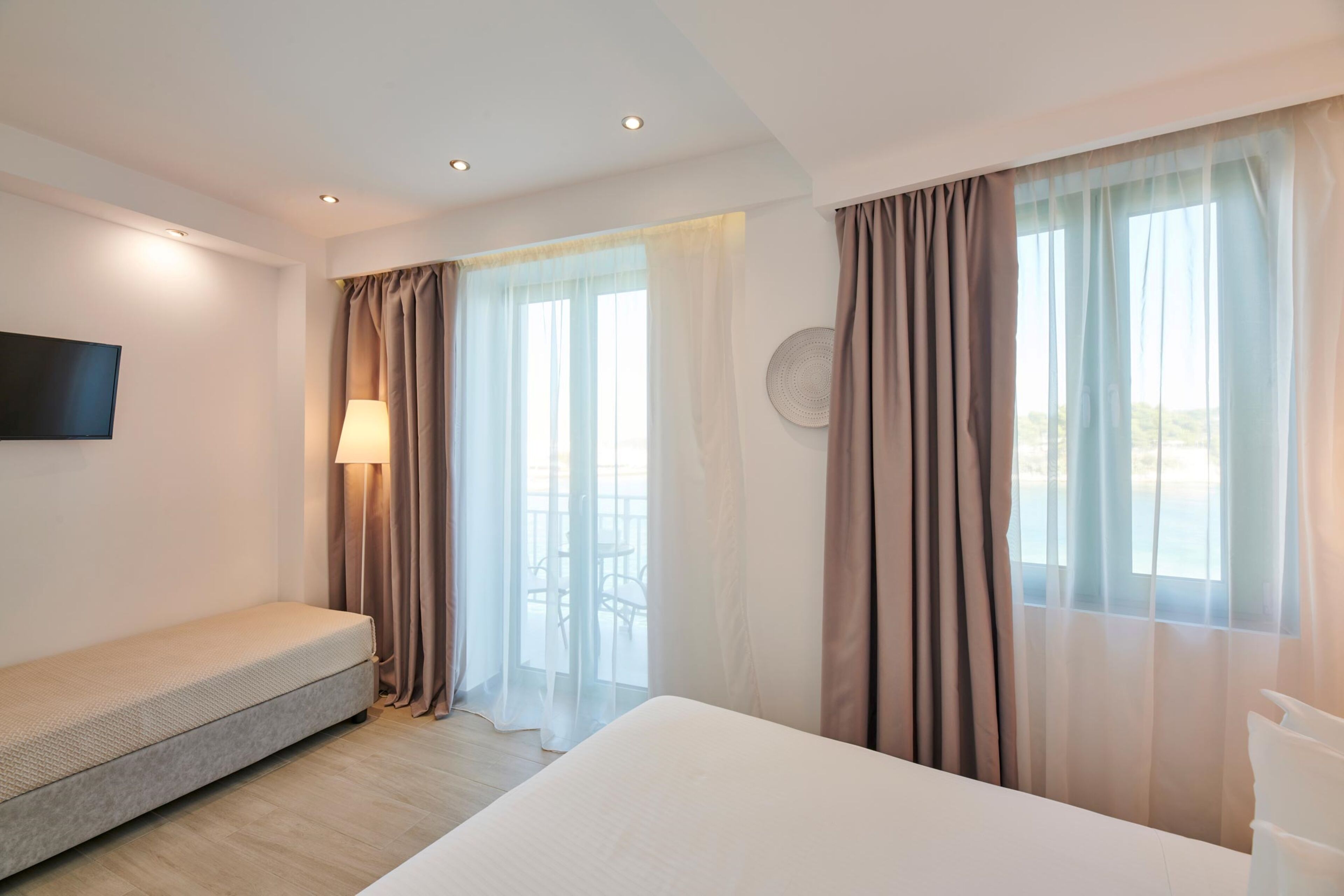 A hotel room with a bed, a sofa, a TV mounted on the wall, and a window with curtains.