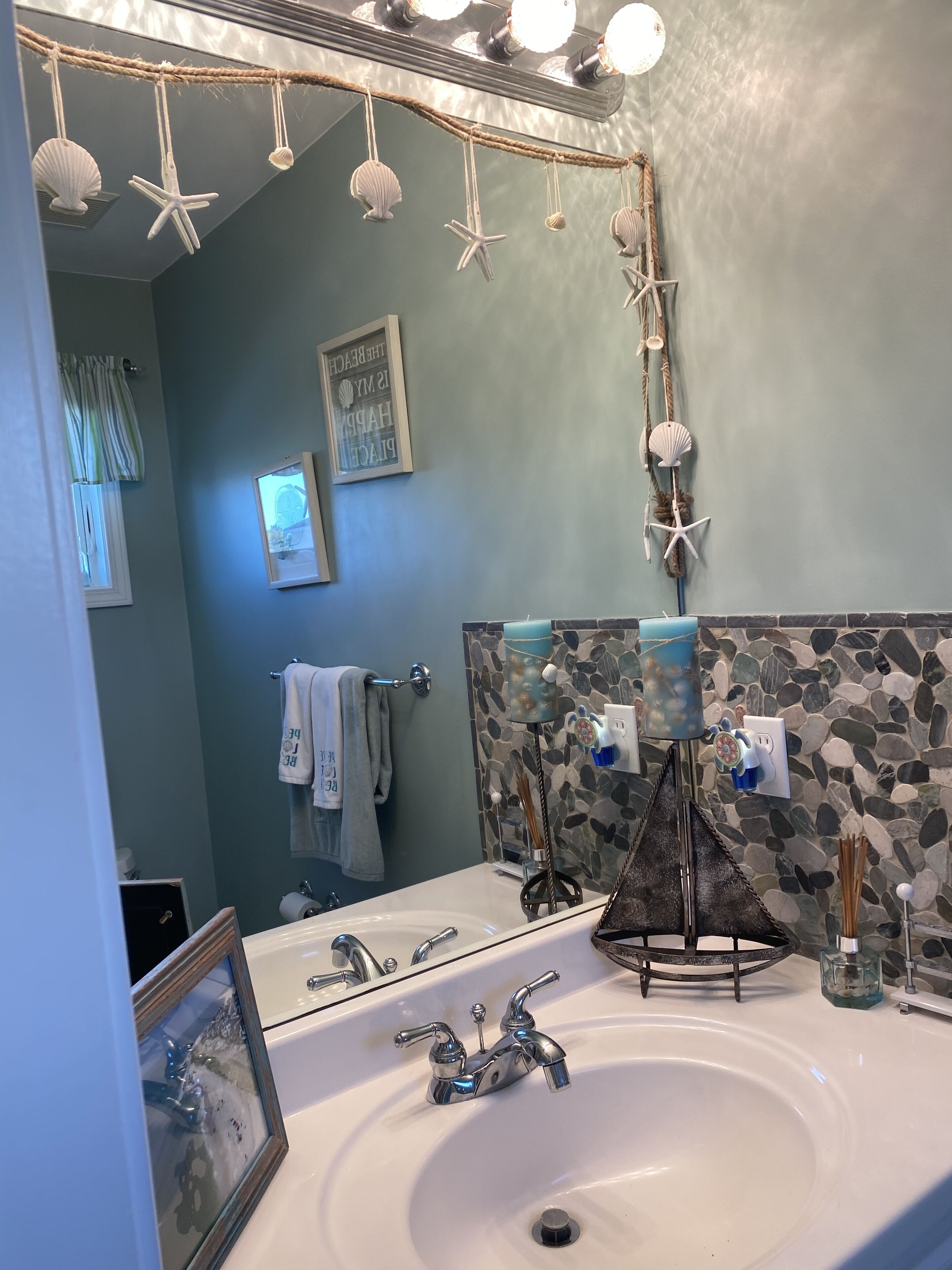 Une salle de bain avec un lavabo blanc, deux robinets, un miroir avec éclairage, des décorations en forme d’étoiles de mer, un tableau encadré et un porte-serviettes.