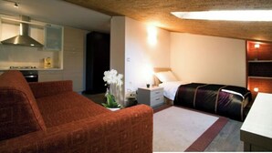 Une chambre d’hôtel compacte comprenant un coin cuisine, un lit recouvert d’une courtepointe noire et violette, un canapé rouge et une petite table blanche.