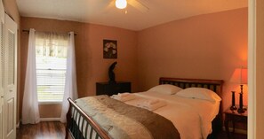 Ein Schlafzimmer mit einem Bett, einem Nachttisch, einer Lampe, einem Gemälde und einem Fenster mit Vorhängen.