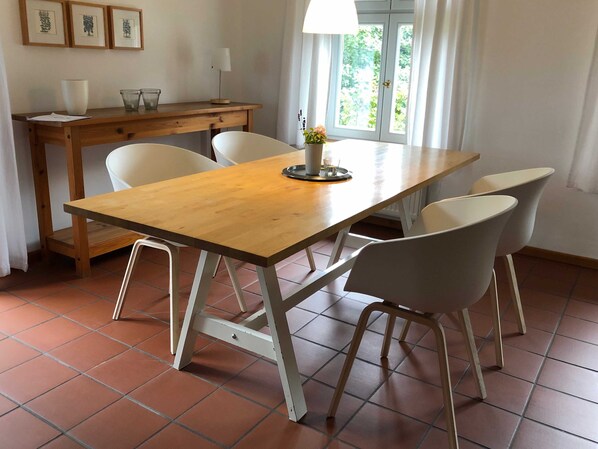 A dining area with a wooden table and four white chairs. There is a small table with a lamp and a vase with flowers. A window with curtains is visible in the background.