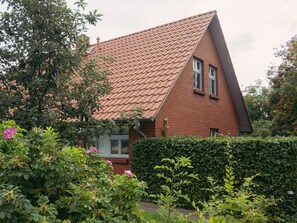 A red brick house with a sloped roof, surrounded by lush greenery and flowering plants.