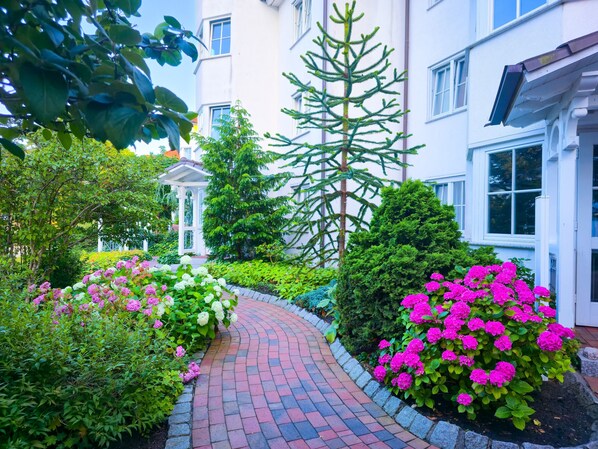 A garden pathway leading to a building with a white facade, surrounded by lush greenery and flowering plants.