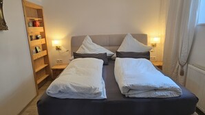 A double bed with pillows, a wooden shelf with books and a lamp, and a nightstand with a lamp.