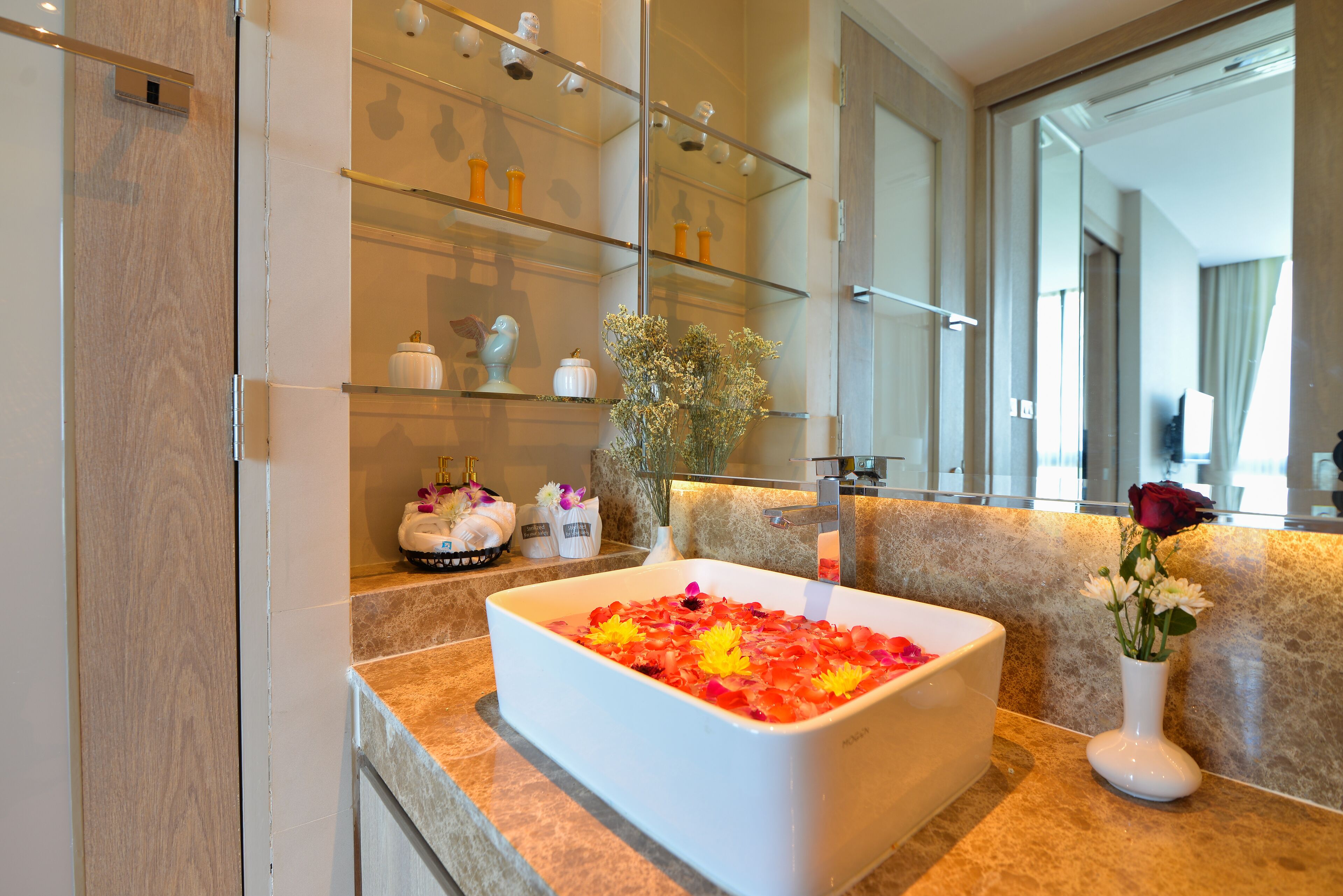 Un baño con una bañera blanca grande llena de pétalos de rosa, un florero blanco con una sola rosa roja y un espejo sobre el lavabo.