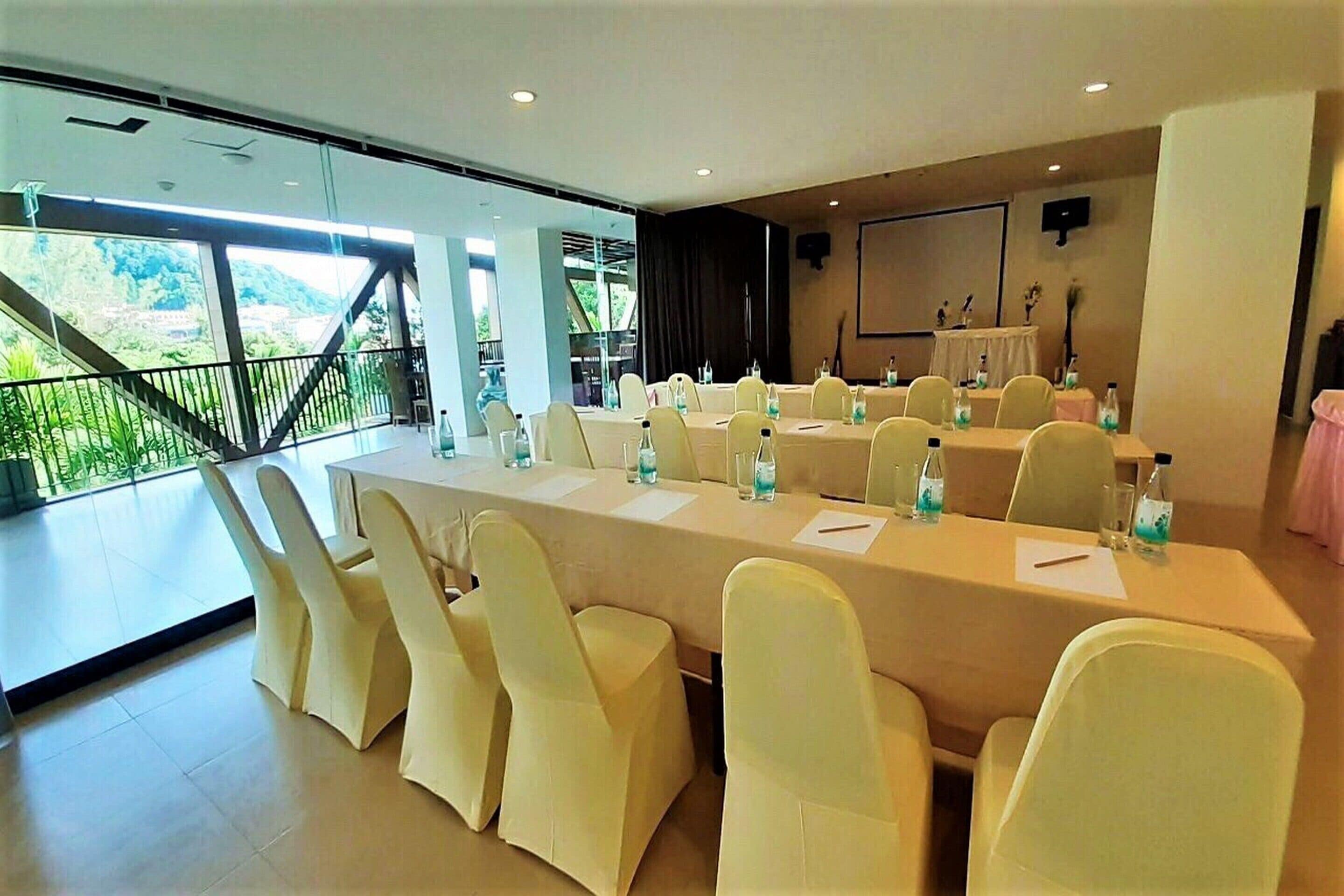A conference room with a long table and chairs, large windows with a view of greenery, and a stage with speakers.