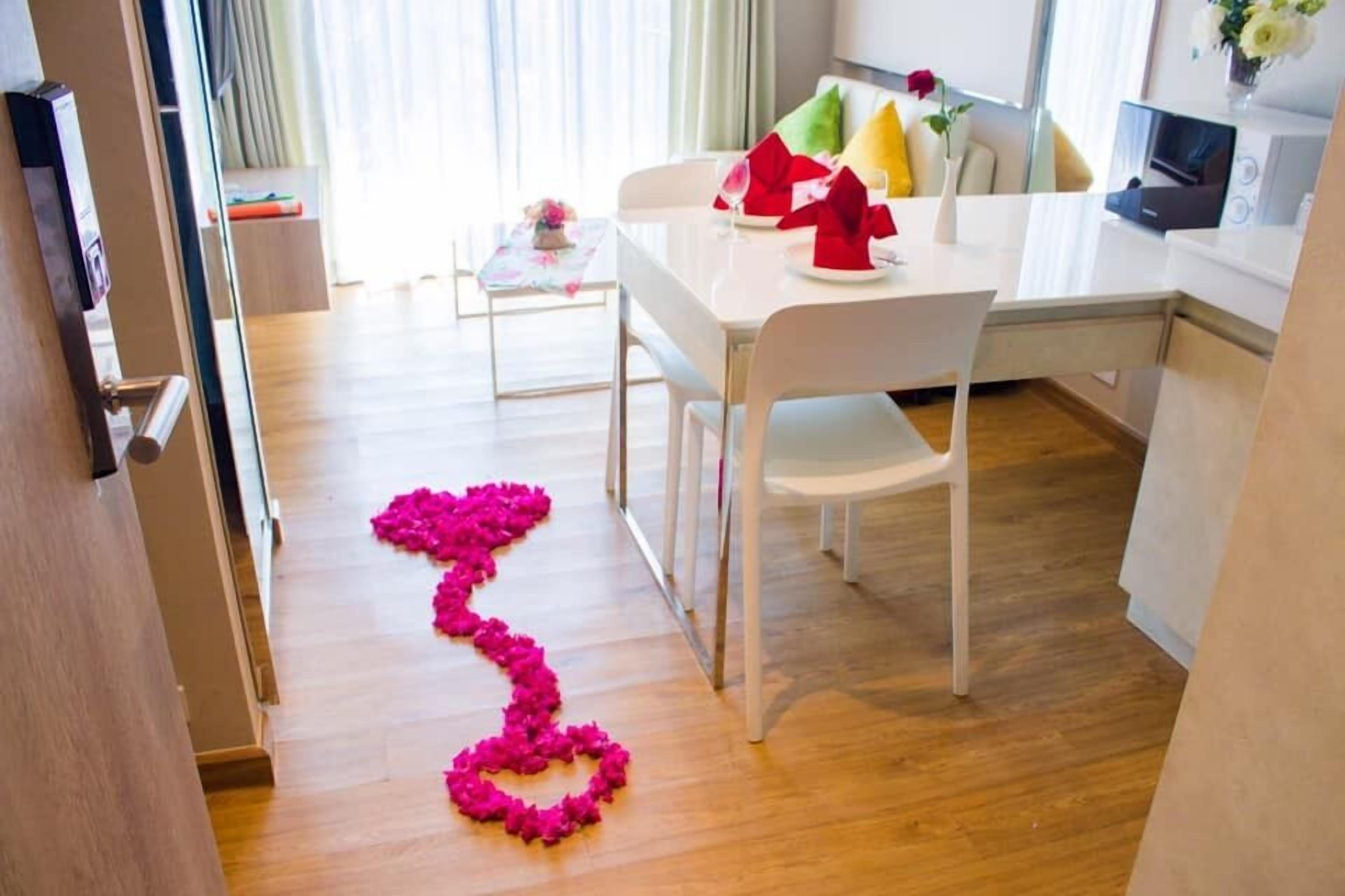 A modern hotel room with a dining area, a sofa with colorful cushions, and a heart-shaped flower arrangement on the floor.