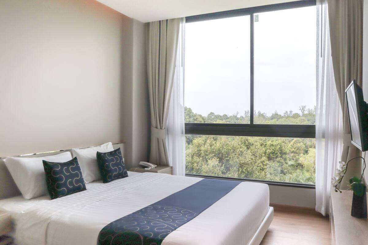 Una habitació d'hotel amb un llit gran, televisió i finestra amb vista a arbres.