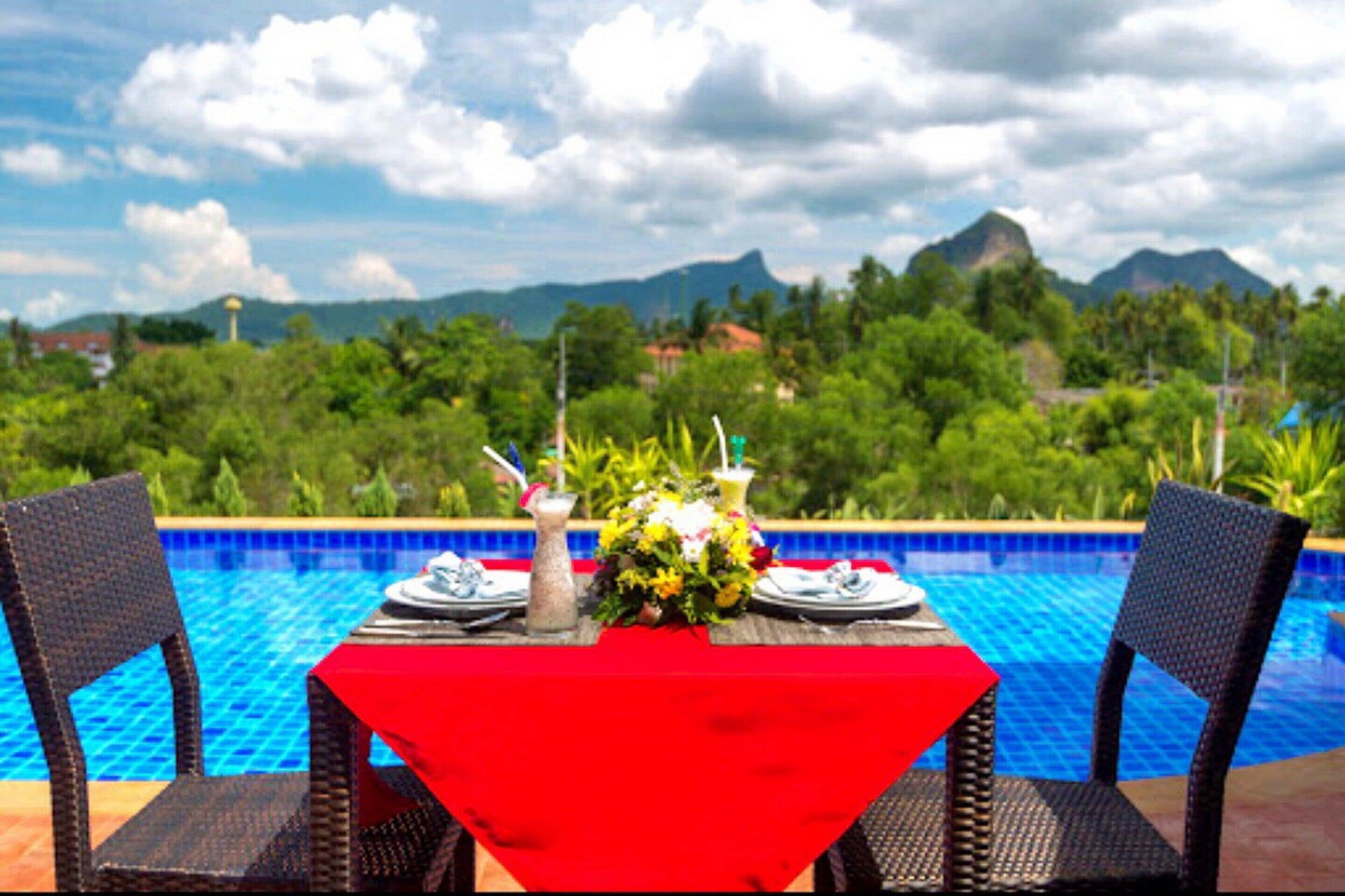 Una mesa de comedor con mantel rojo, dos sillas de mimbre y vistas a una piscina y montañas al fondo.