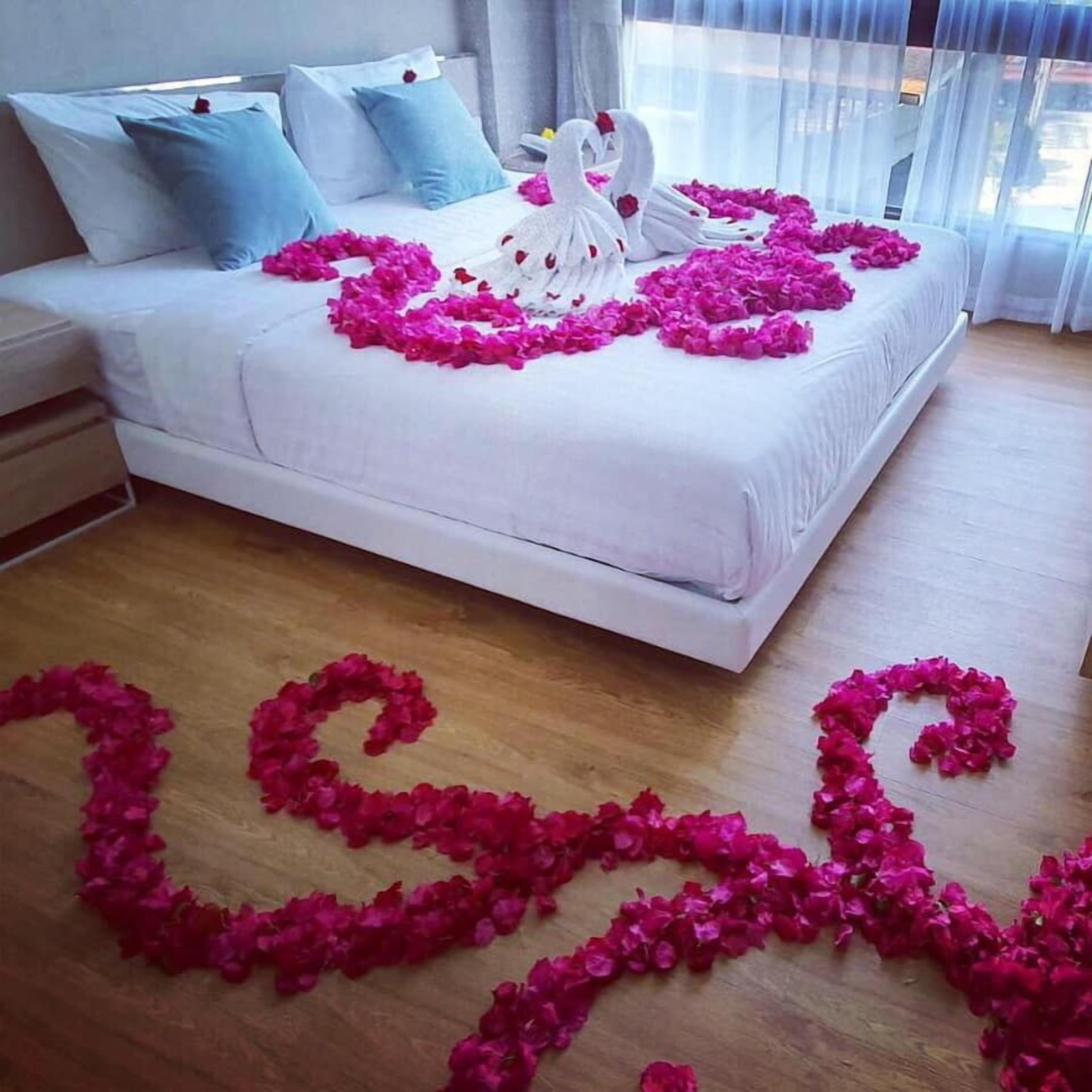 A bed with white linens, pink rose petals arranged in a heart shape on the floor, and two swan-shaped pillows.