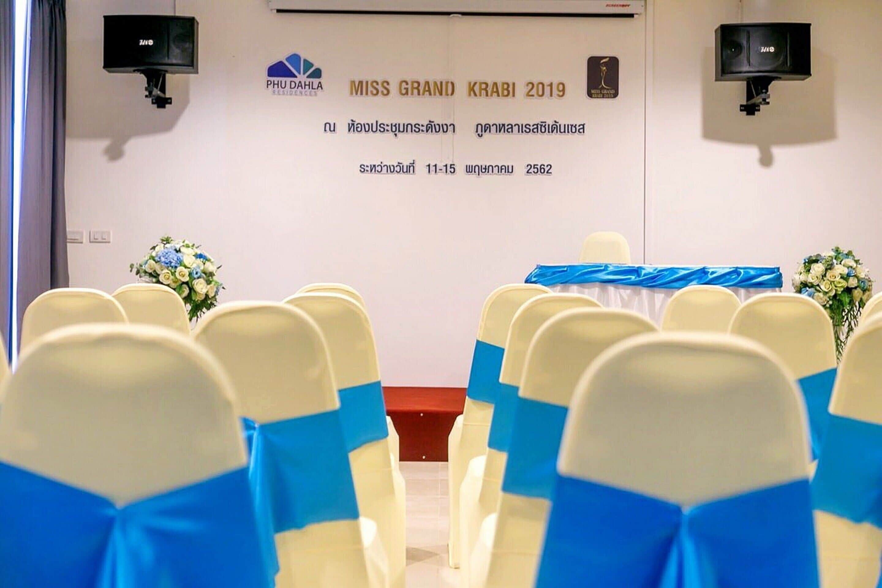 A room with rows of chairs, a stage with a banner, and two wall-mounted speakers.