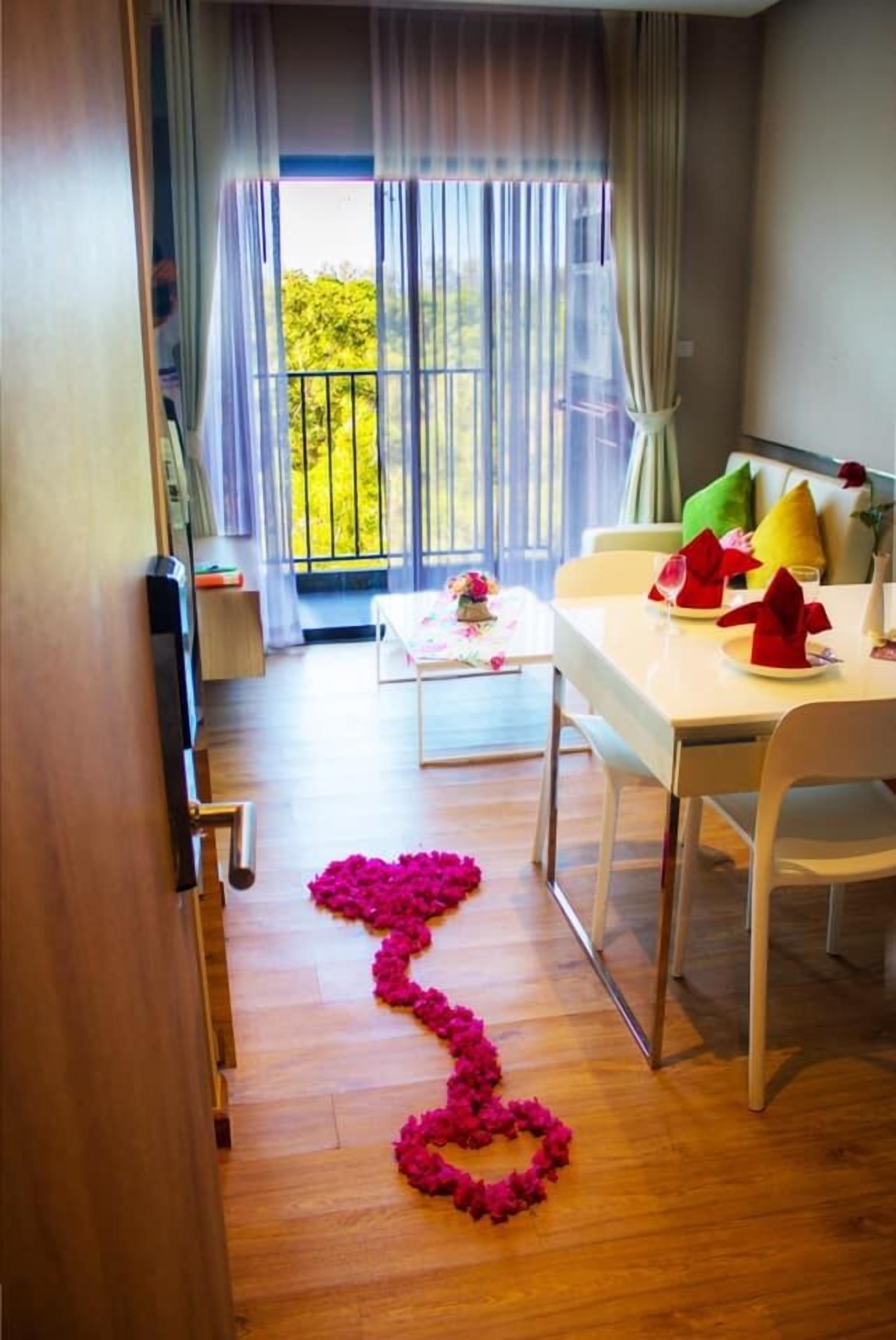 Una habitación con una mesa de comedor para dos, una mesita con flores y un balcón con vistas al verdor.