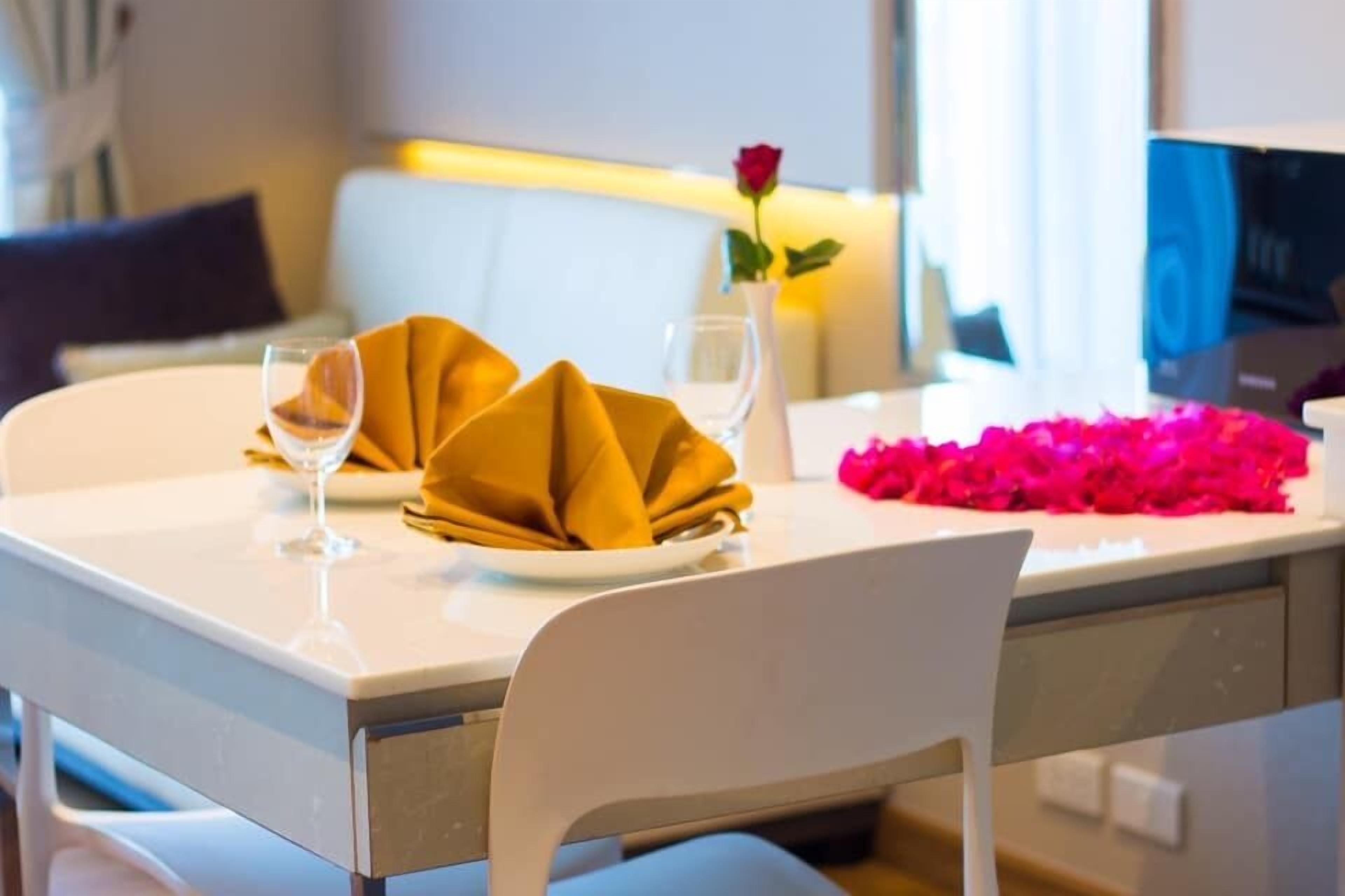 A dining table with a white tablecloth, two folded napkins, two wine glasses, a vase with a single rose, and a pink flower arrangement.