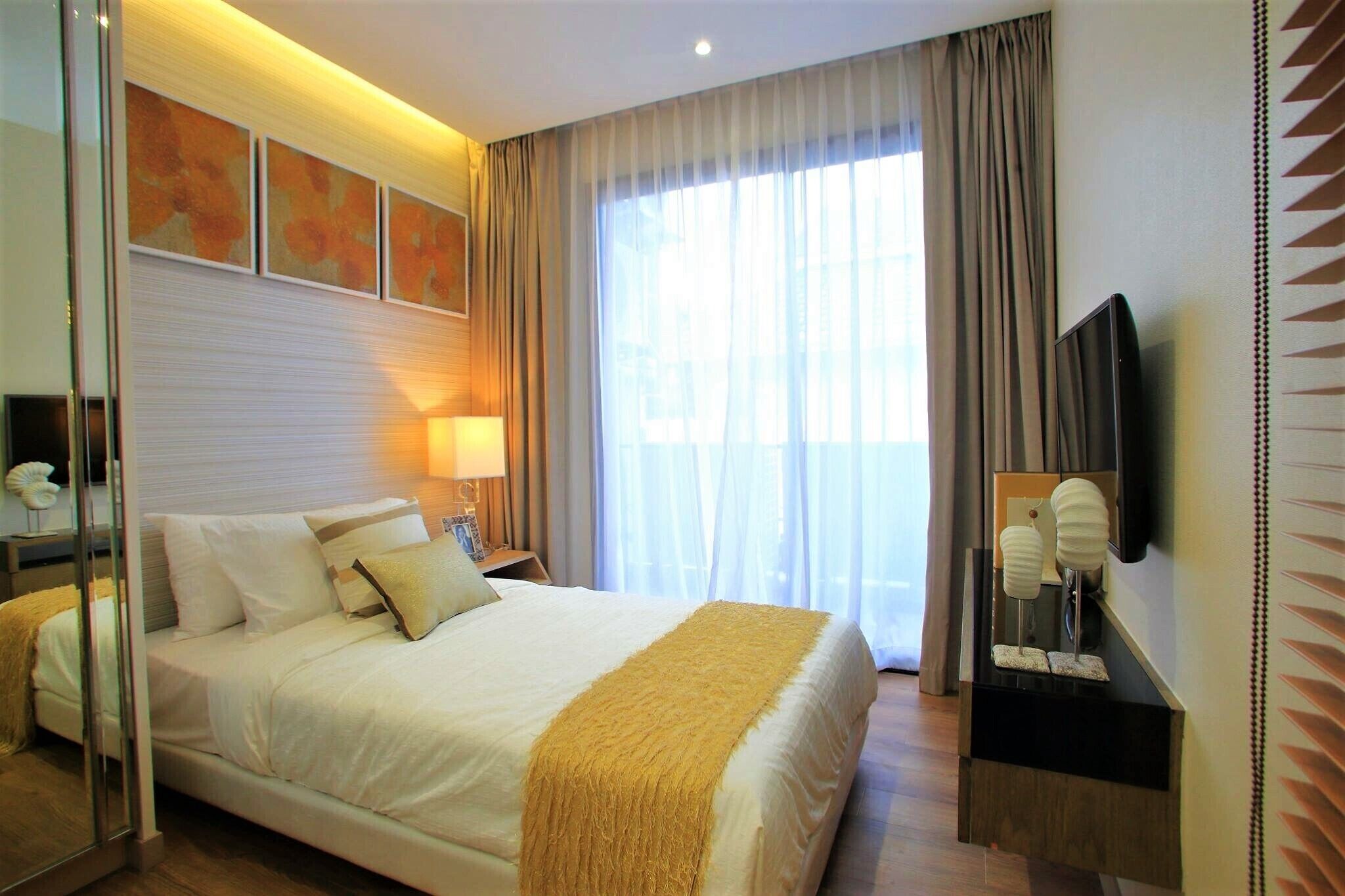 A hotel room with a large bed, a bedside table with a lamp, a TV mounted on the wall, and a window with curtains.