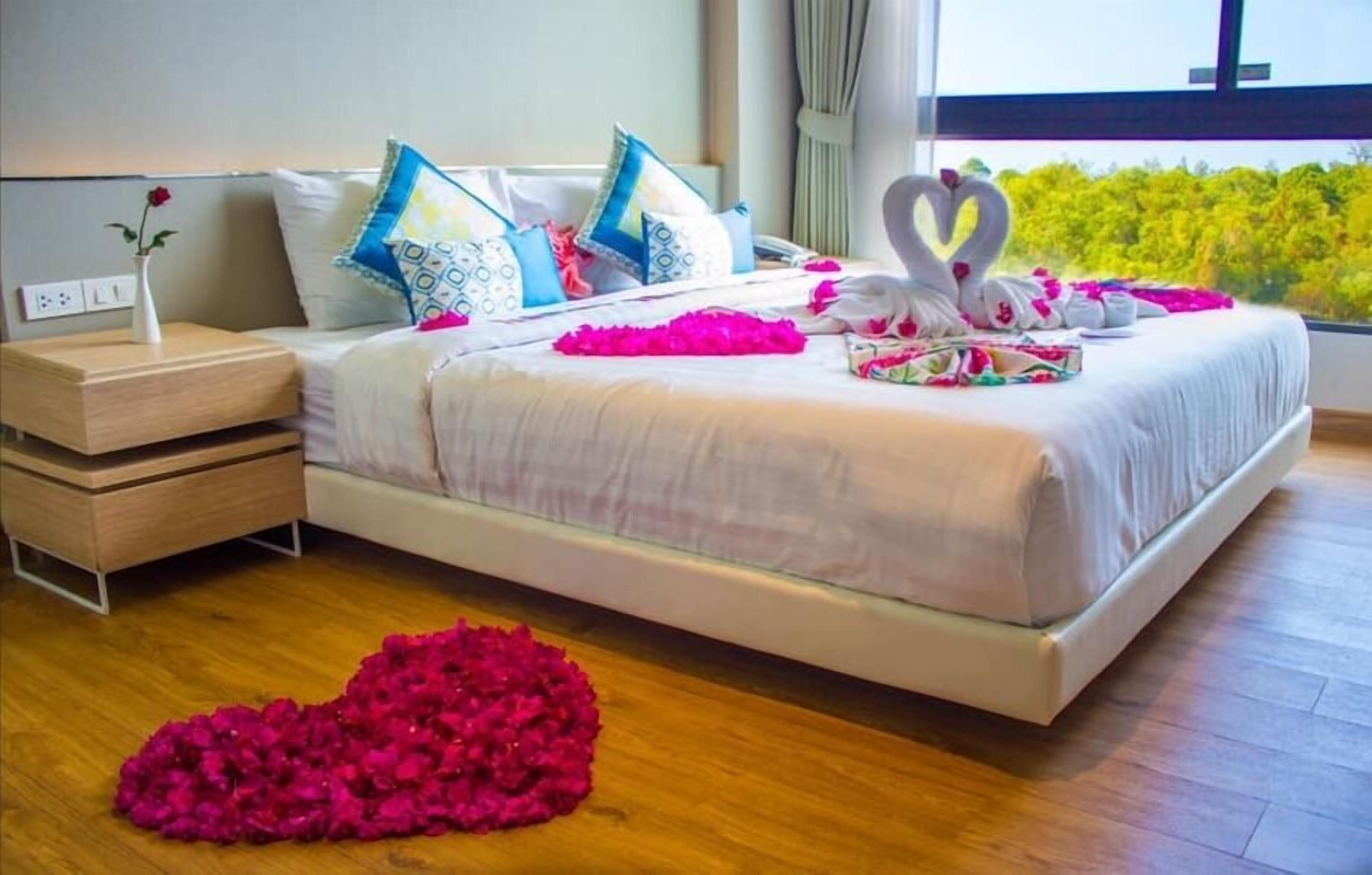 A neatly made bed with a heart-shaped arrangement of rose petals, a bedside table with a vase, and a window with a view of greenery.