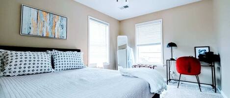 A bedroom with a large bed, two windows, a ceiling fan, a red chair, and a desk with a lamp.