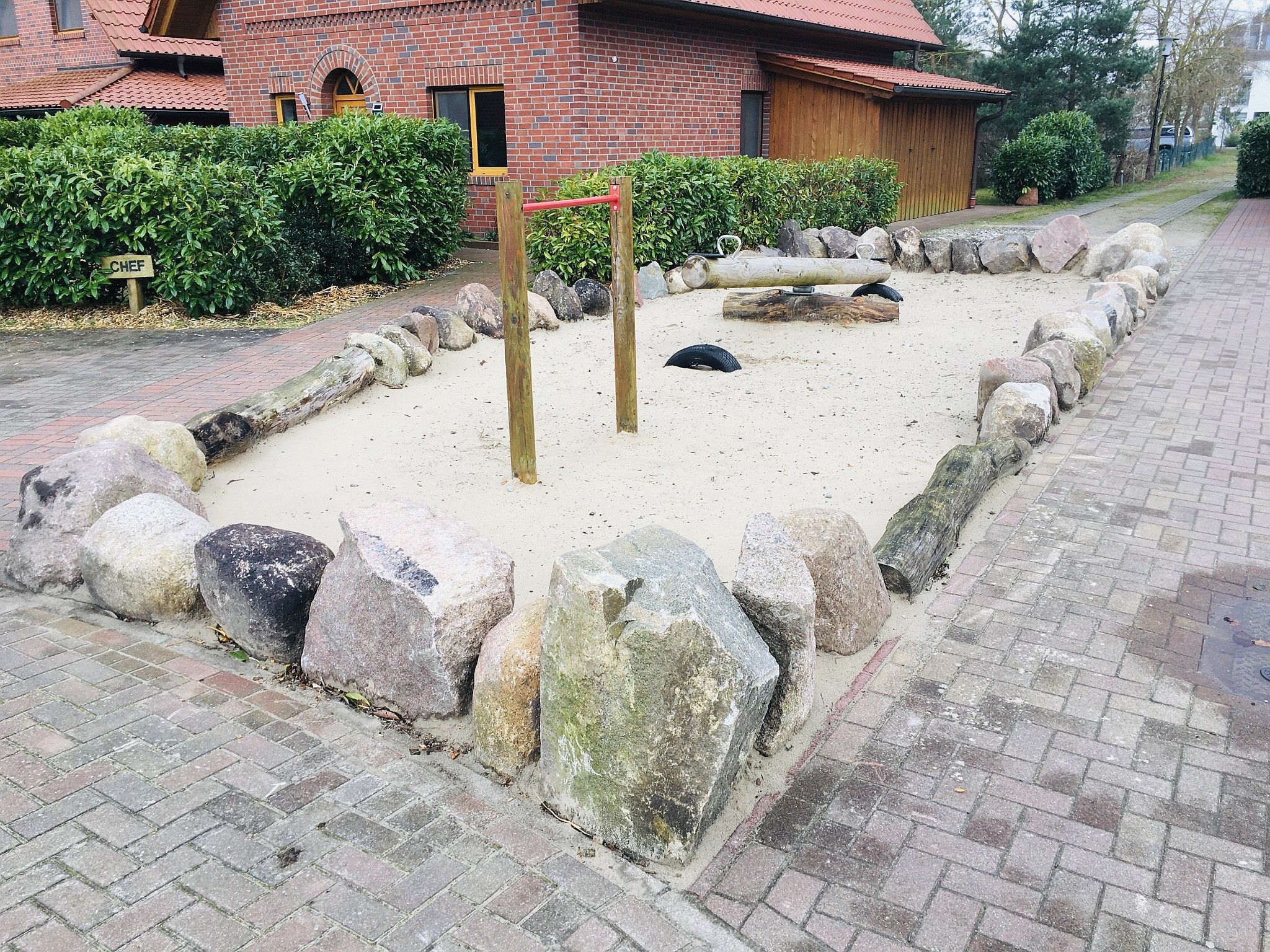 Zona de juegos arenosa con rocas grandes y un conjunto de columpios de madera.