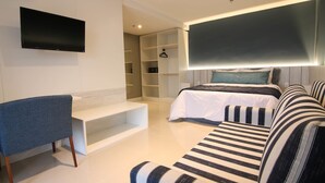 Ein modernes Hotelzimmer mit Flachbildfernseher an der Wand, einer Couch mit gestreiften Kissen, einem Bett mit Kopfteil und einer eingebauten Regaleinheit.