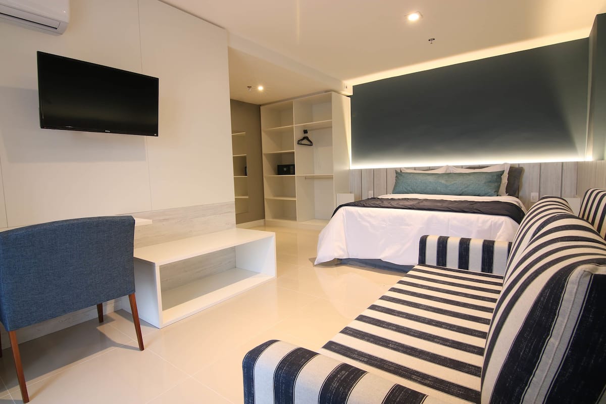 A modern hotel room with a flat-screen TV mounted on the wall, a sofa with striped cushions, a bed with a headboard, and a built-in shelving unit.