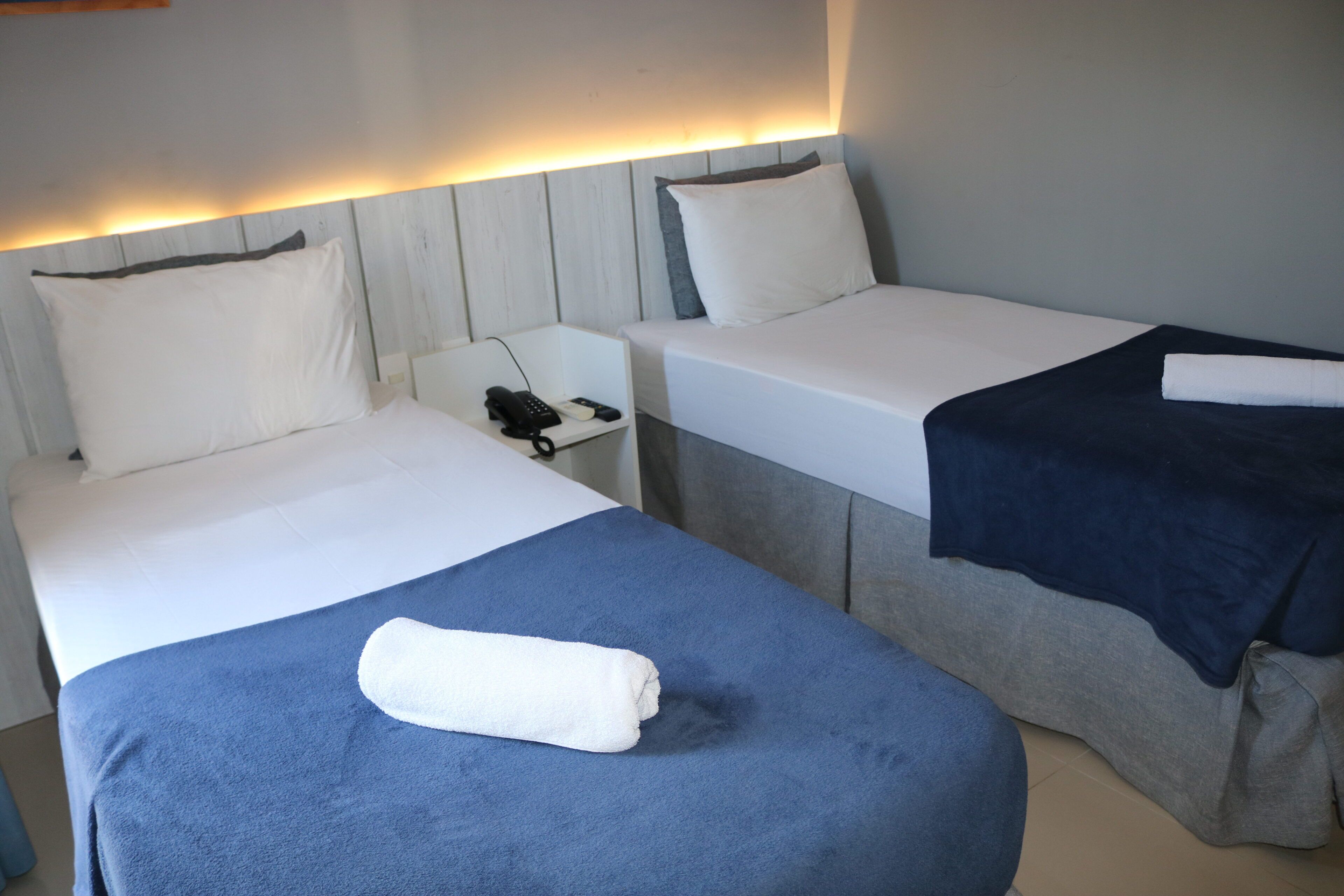 Habitación de hotel con dos camas, cada una con almohada y manta. Se ve una mesita de noche con un teléfono y un control remoto.