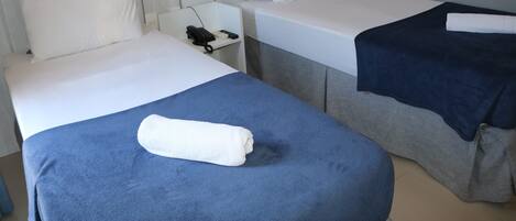 Habitación de hotel con dos camas, cada una con almohada y manta. Se ve una mesita de noche con un teléfono y un control remoto.