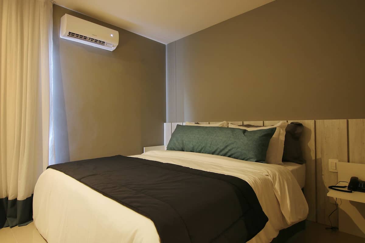 A hotel room with a bed, a bedside table, a wall-mounted air conditioner, and a window with curtains.