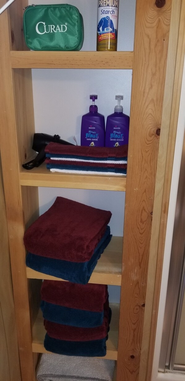 Ein Holzregal mit einer grünen Tasche mit dem Aufdruck 'GURAD', zwei lila Flaschen 'Aussie Miracle Moist Shampoo' und mehreren gefalteten Handtüchern in Rot und Blau.