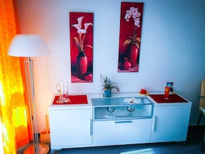 Une pièce avec un meuble blanc, une table à plateau en verre, un vase rouge et deux tableaux rouges accrochés au mur.