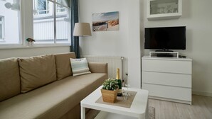 A modern living room with a beige sofa, a white coffee table with a plant, a TV on a white stand, and a window with curtains. - 15 Ferienappartement Mönchgut (A) - Apartment house Haus Arkona (Sellin)