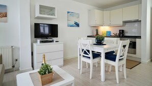 A modern kitchen with white cabinets, a dining table with blue cushions, and a TV on a white stand. - 15 Ferienappartement Mönchgut (A) - Apartment house Haus Arkona (Sellin)