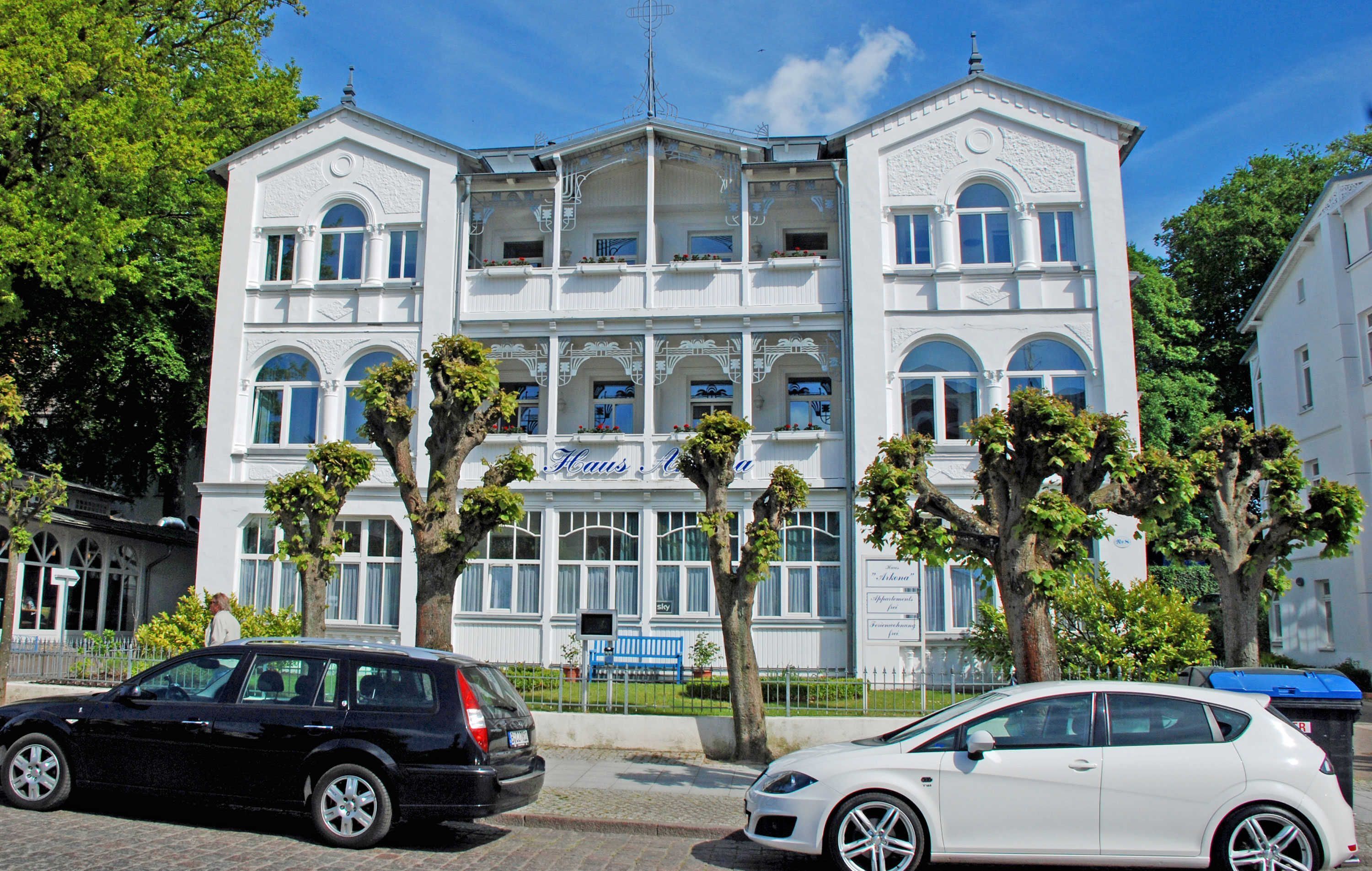 Un edificio de varios pisos con una entrada central y balcones, rodeado de árboles y coches estacionados.