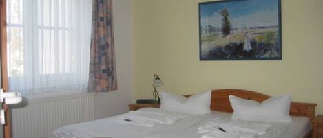 A bedroom with a wooden bed, white bedding, a painting on the wall, and a window with sheer curtains.