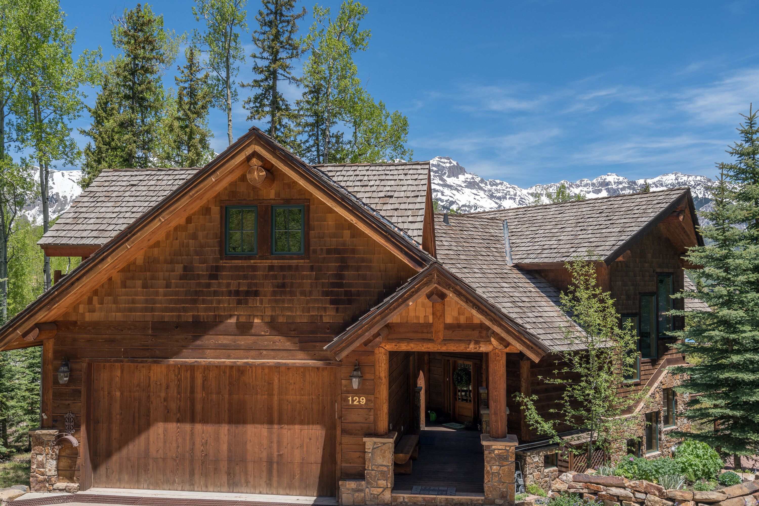 A wooden cabin with a stone entrance, a number 129 sign, and a view of snow-capped mountains.