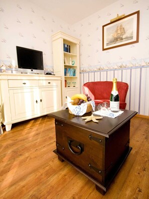 Une pièce comprenant un coffre en bois, un fauteuil rouge, une bibliothèque, un tableau encadré accroché au mur et un téléviseur posé sur un meuble.
