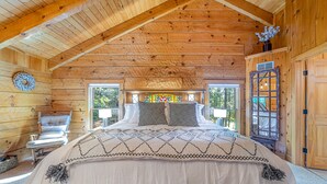 A log cabin bedroom with a large bed, a chair, and a window with a view of trees. - Luxury cabin in the woods, 2 bedrooms plus loft (Fairfield)