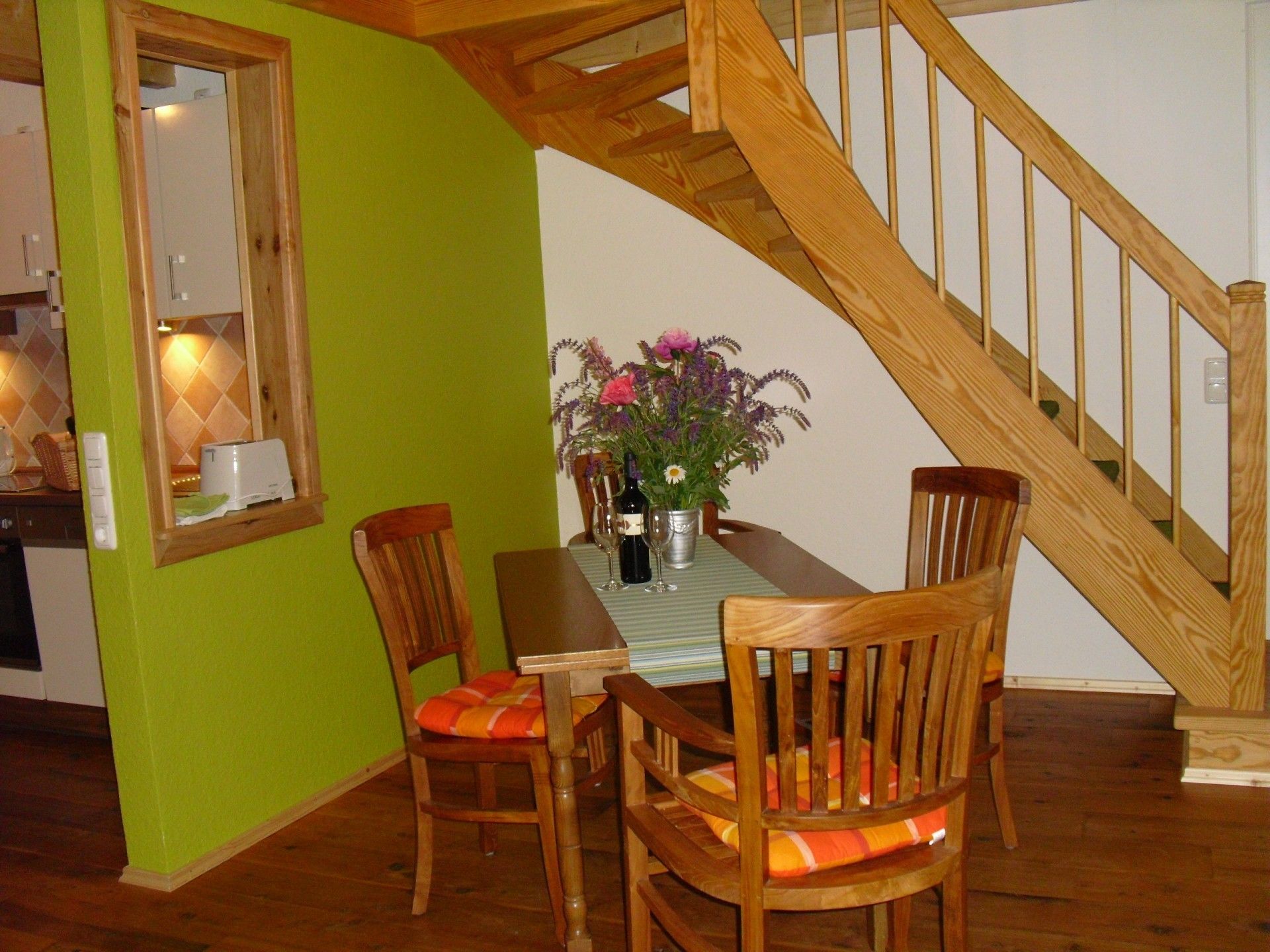 A dining area with a wooden staircase, green walls, and wooden chairs with orange cushions.